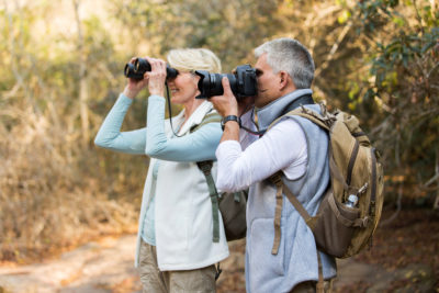 An image of an older couple looking towards the left; the girl is behind the boy and has binoculars, the boy is using a camera.