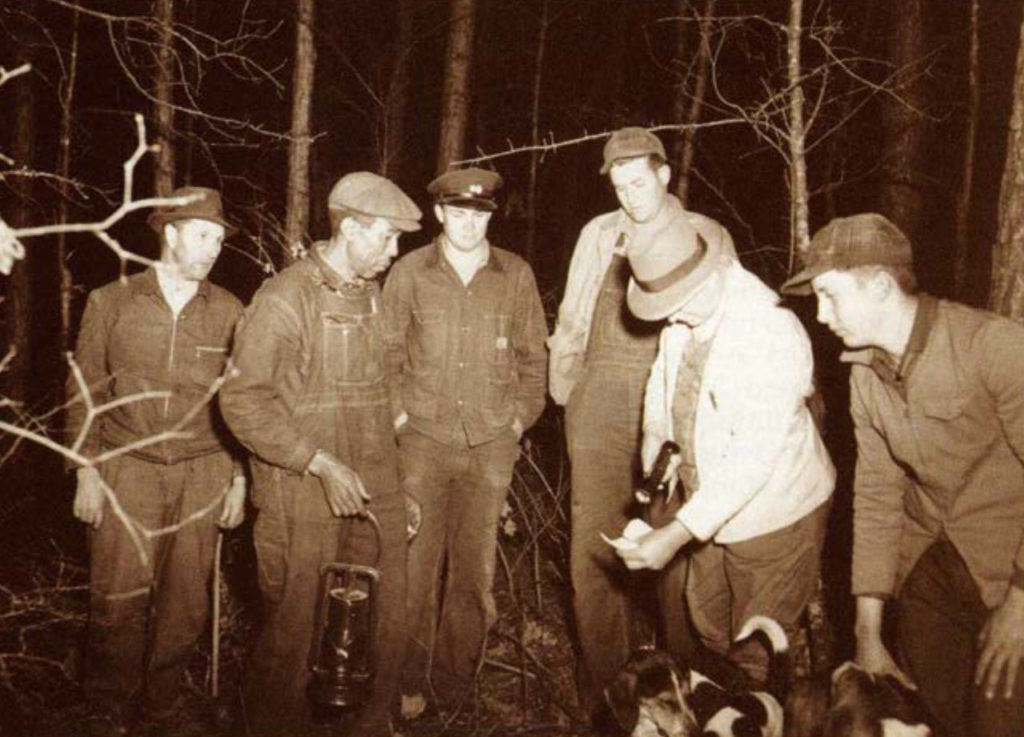A black and white picture of two game wardens checking the hunting licenses of a group of raccoon hunters