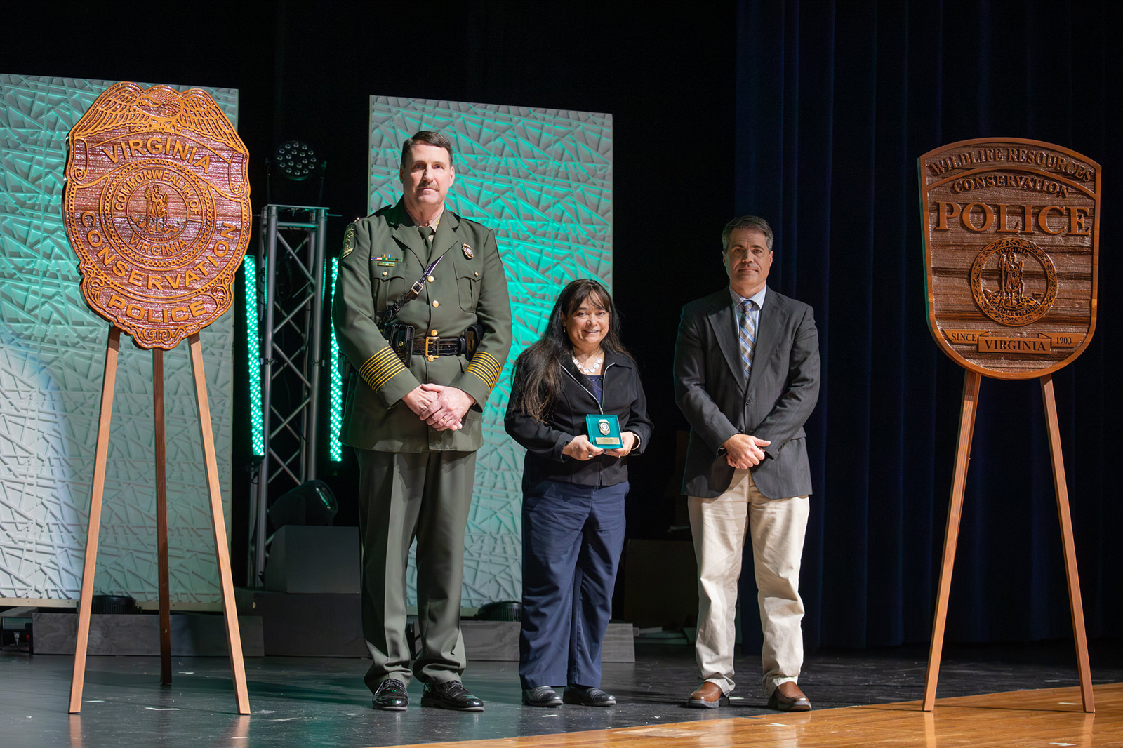 A photo of a woman holding an award, standing on a stage with a man in Conservation Police uniform and another man.