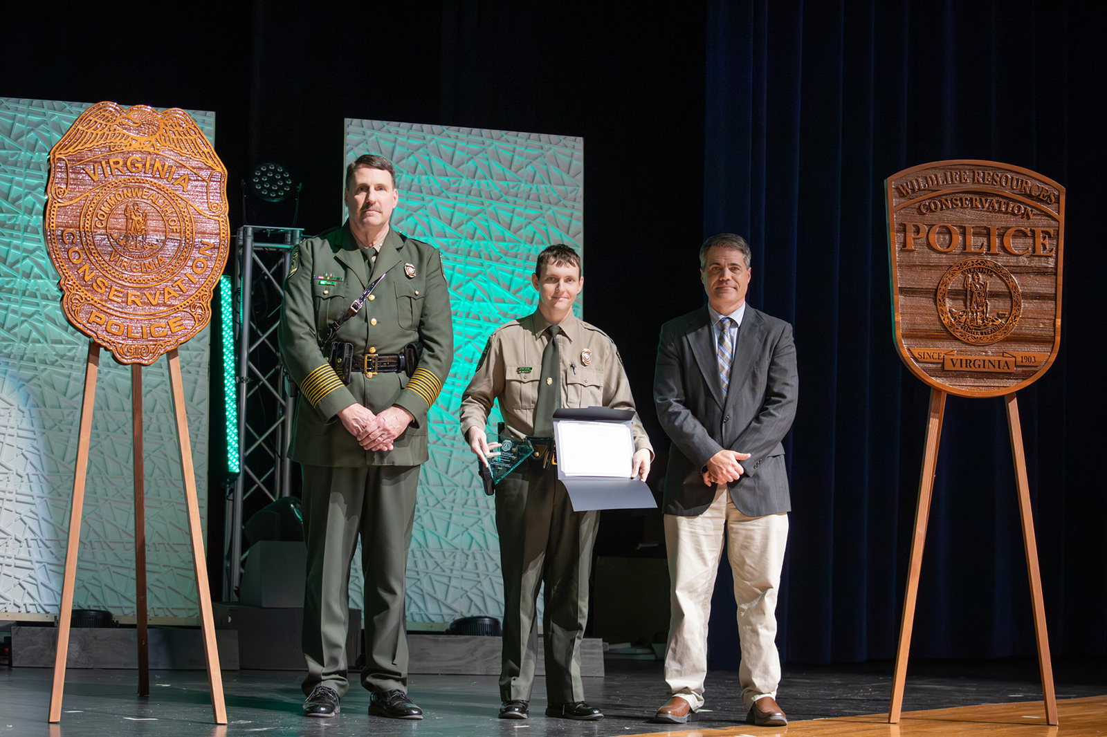 A photo of a Conservation Police Officer in uniform holding an award standing on a stage between another CPO in uniform and a man in a blazer.