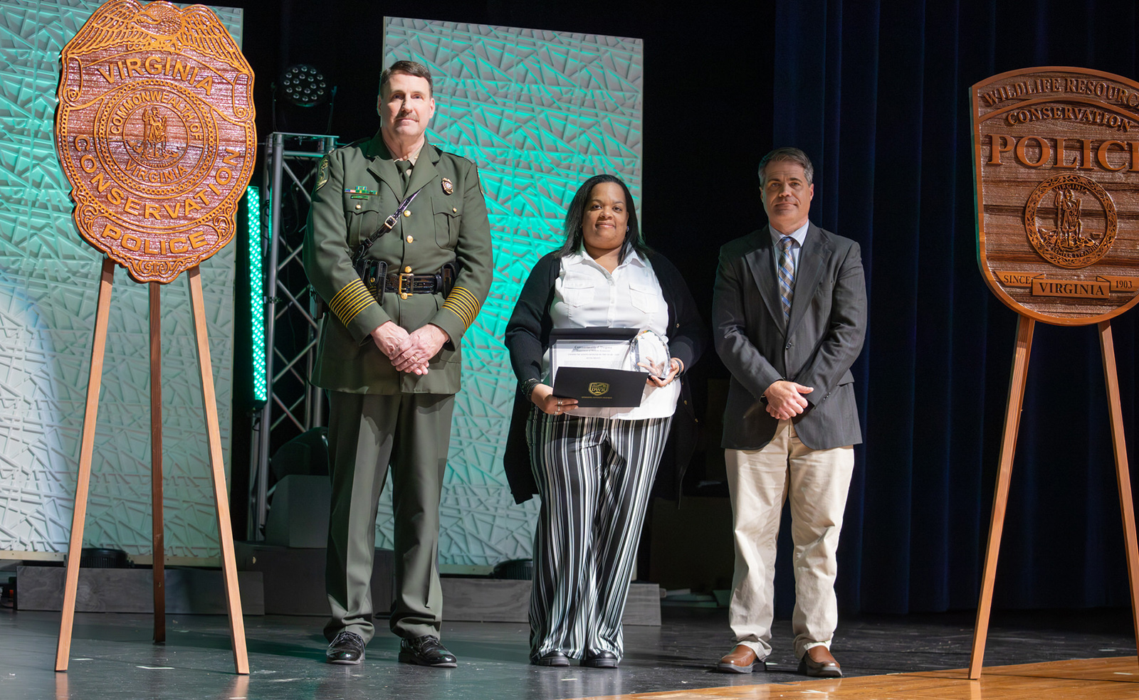 A photo of a woman holding an award, standing on a stage with a man in Conservation Police uniform and another man.