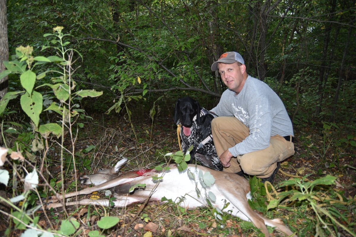 An image of a German Shorthaired Pointer sitting next to his owner in a deciduous forest; in front of them is a dead Botetourt doe. 