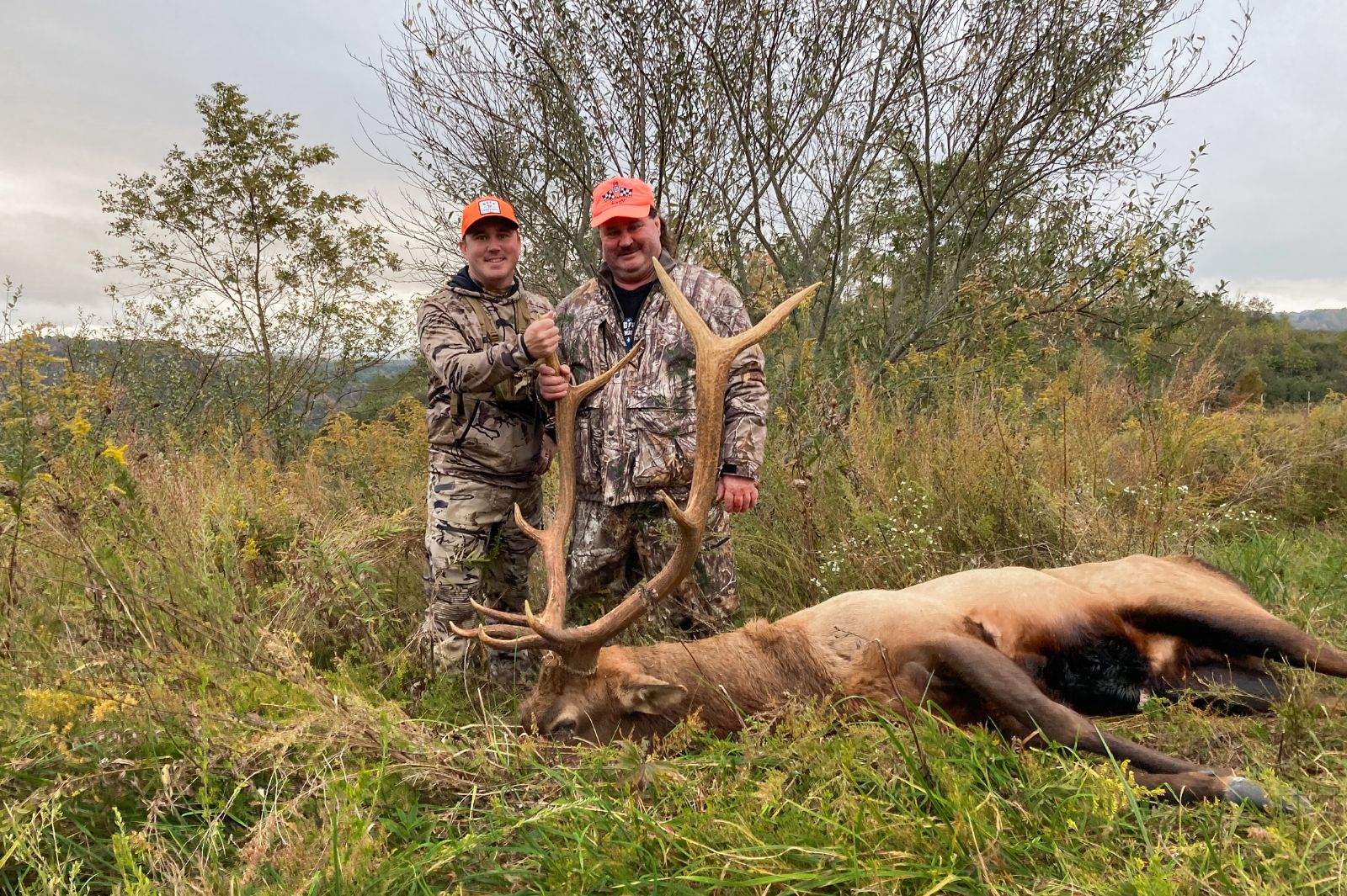 A photo of two hunters in camoflague and blaze orange posing with a large bull elk lying on the ground.