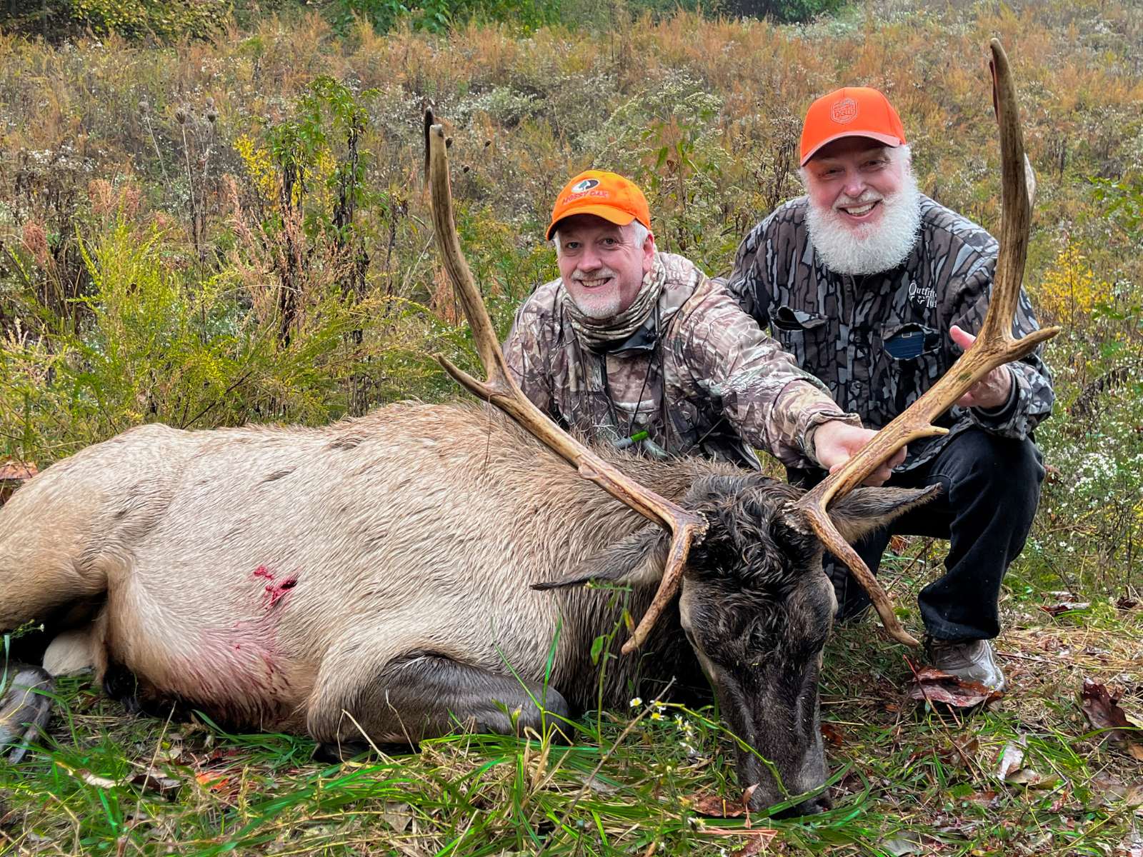 A photo taken in the nighttime of two hunters in camoflague and blaze orange posing with a large bull elk lying on the ground.