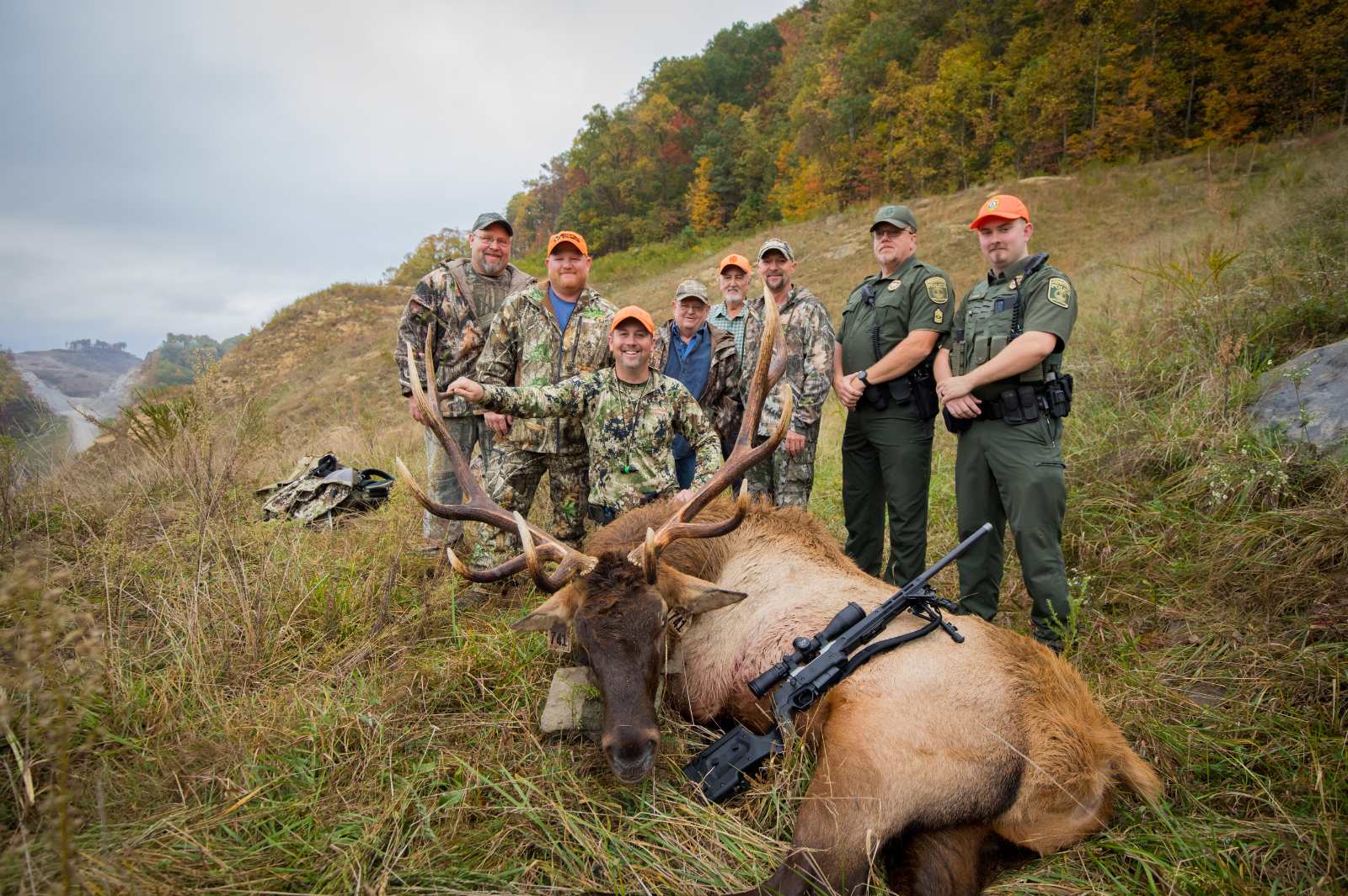 A photo taken of six hunters in camoflague and blaze orange posing with a large bull elk lying on the ground and two Conservation Police Officers in uniform.