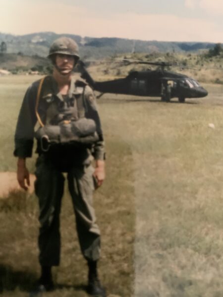 An older photo of a man in military uniform standing in a field with a Blackhawk helicopter in the background.