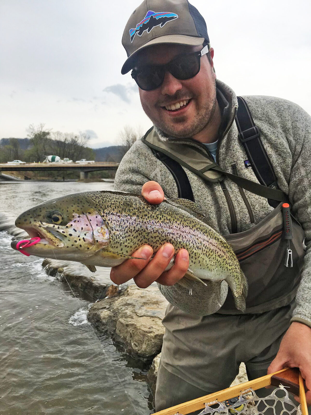 A man smiles at the camera while holding a rainbow trout in one hand.