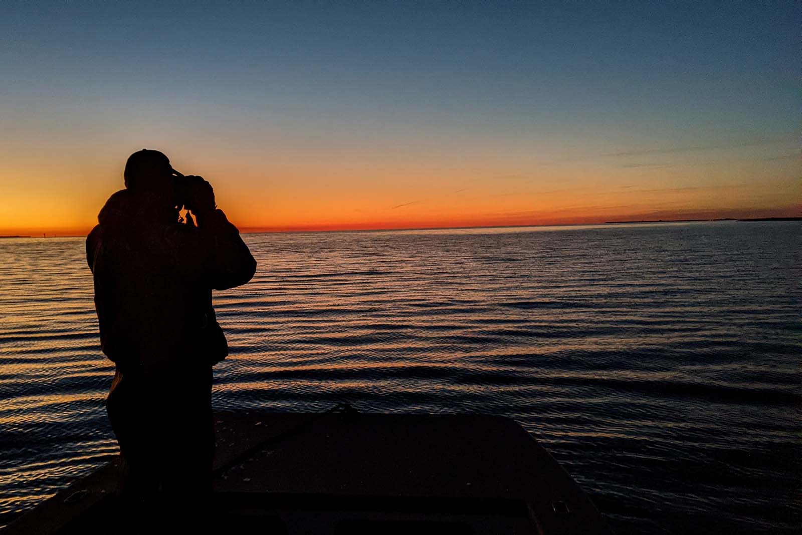 A Conservation Police Officer standing on a platform at dawn and looking through binoculars out over the water