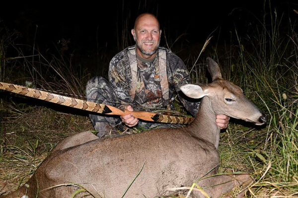An image of a hunter with his bow an arrow and a doe he has killed