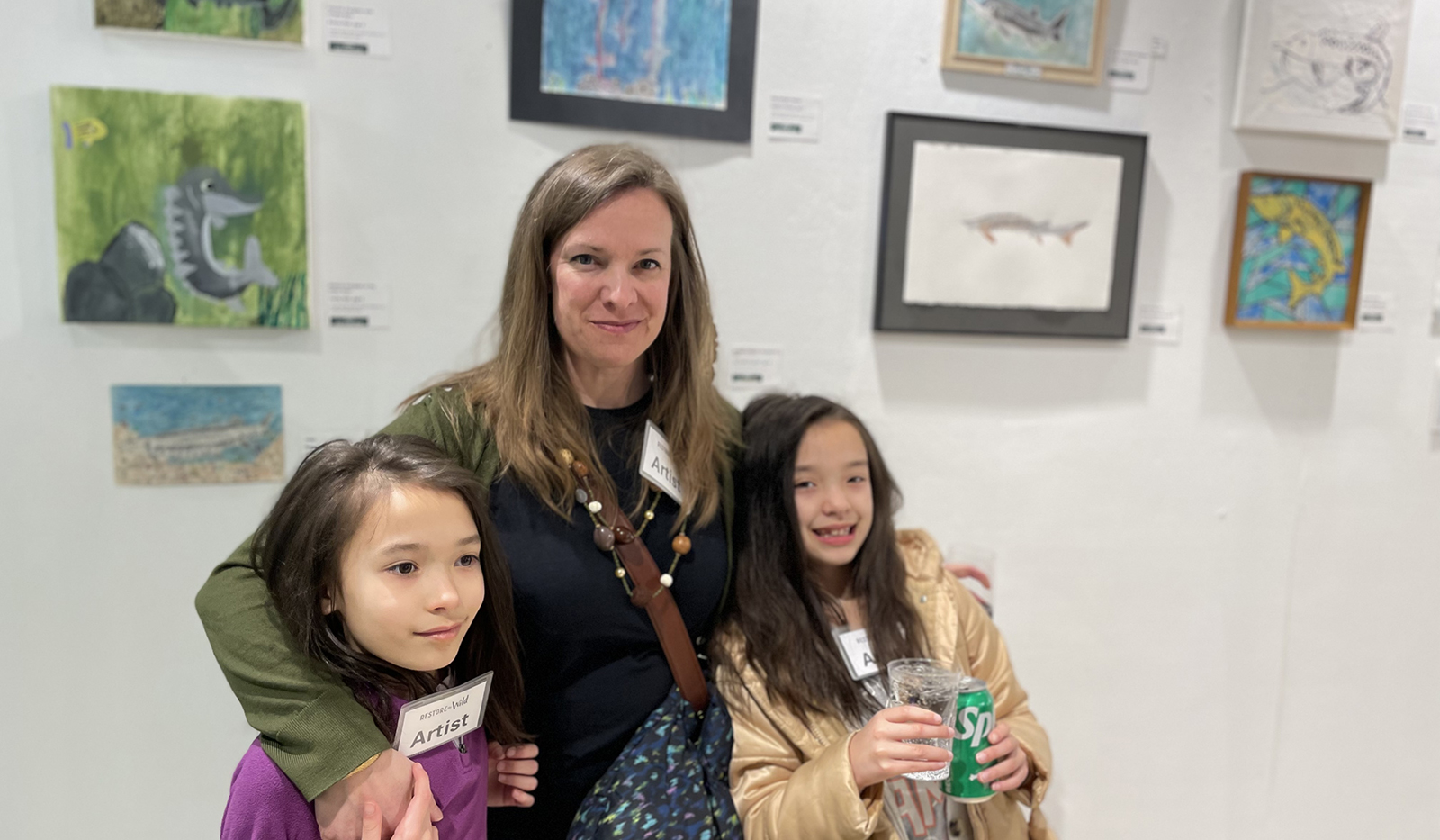 A photo of a woman and two 8-year-old girls standing in front of a wall full of paintings.