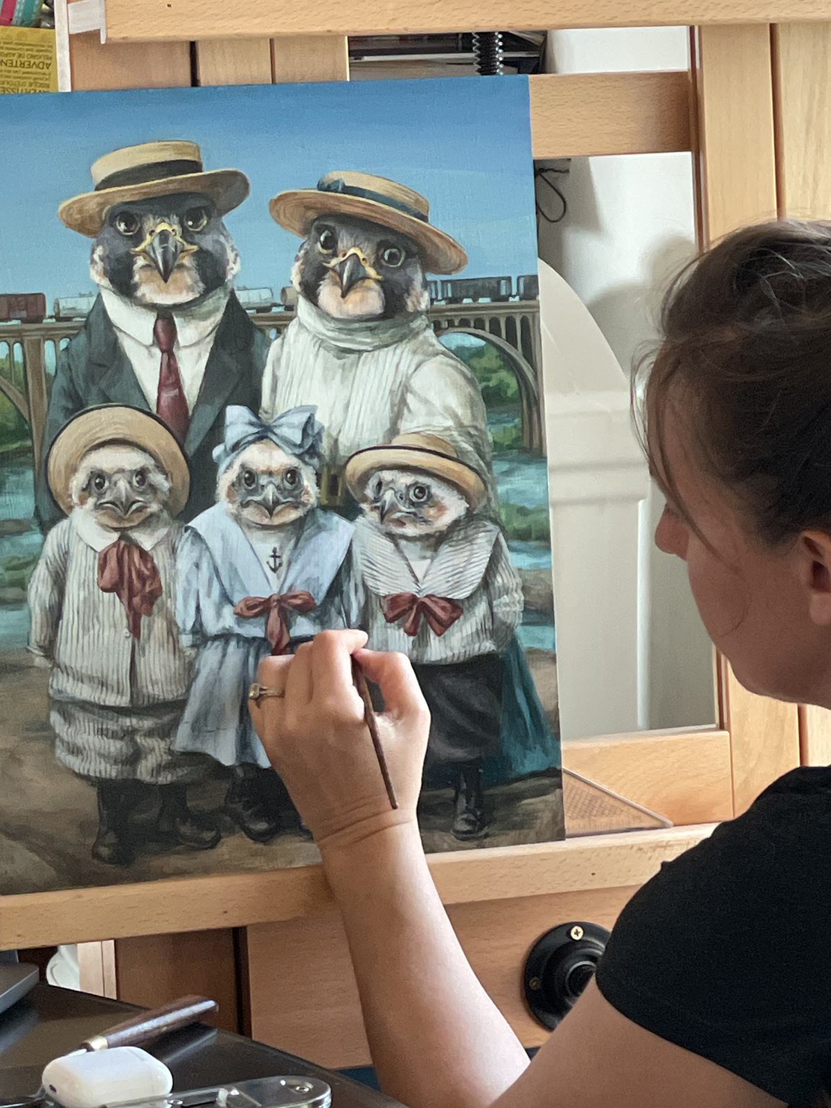A photo of a woman painting a large painting of two adult and three juvenile peregrine falcons dressed in clothes in front of the James River in Richmond.