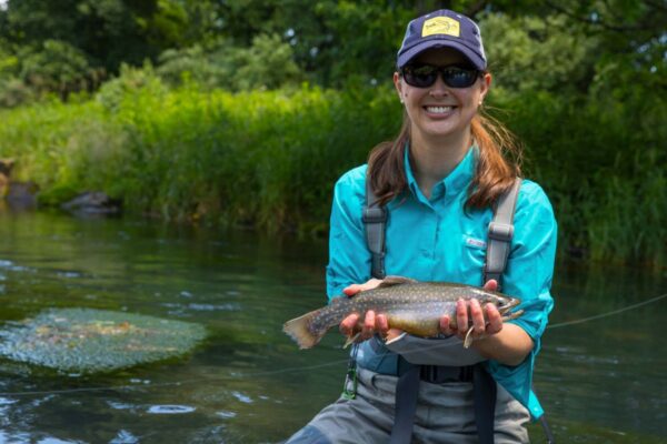 Woman in fishing gear, smiling and holding a brook trout for the picture.