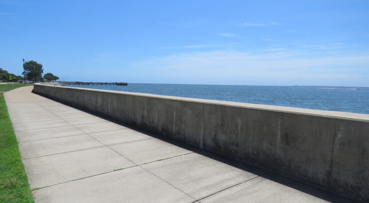 An image of Fort Monroe's seawall, it is in a grassy field with a pier in the distance; the wall itself is concrete with a sidewalk next to it
