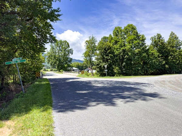A scenic view of a rural intersection with a gravel road branching off to the right. Lush green trees line both sides of the road under a clear blue sky, with a few clouds visible. A street sign indicates the direction of a nearby road, and a distant view of mountains can be seen in the background.