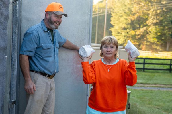 A man in a blaze orange hat and a women in a blaze orange sweater. The woman is holding two wrapped packages of venison while the man looks on, smiling.