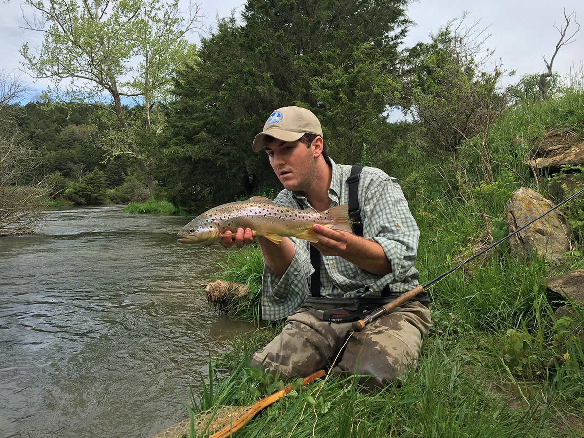 A man kneeling in the grass by the water while looking at a trout he's holding.