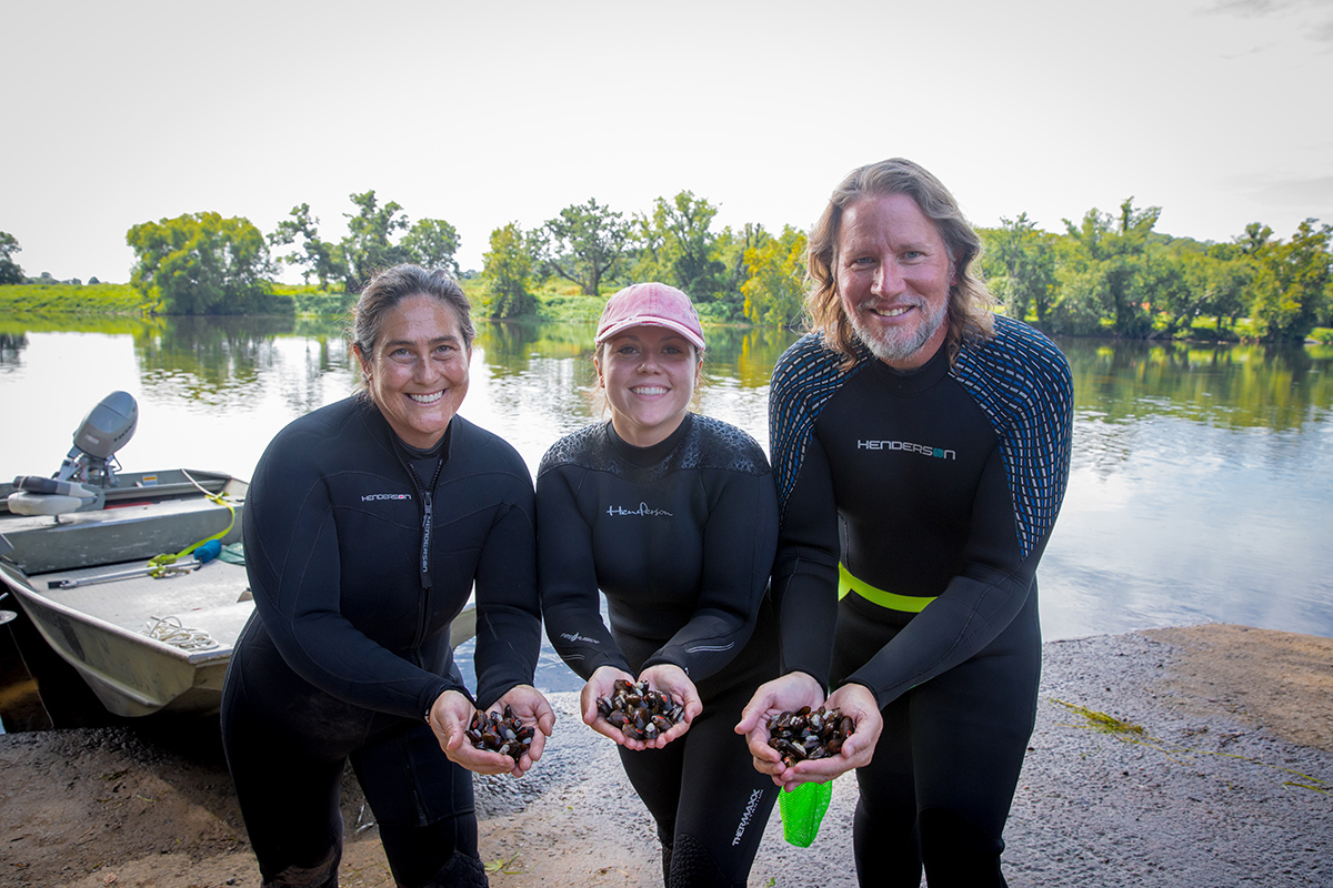 An image of three DWR employees holding handfuls of mussels with a boat and the James river in the background