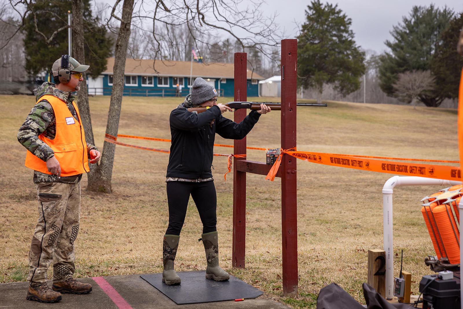 A photo of a man wearing camouflage and orange observing a woman target shooting.