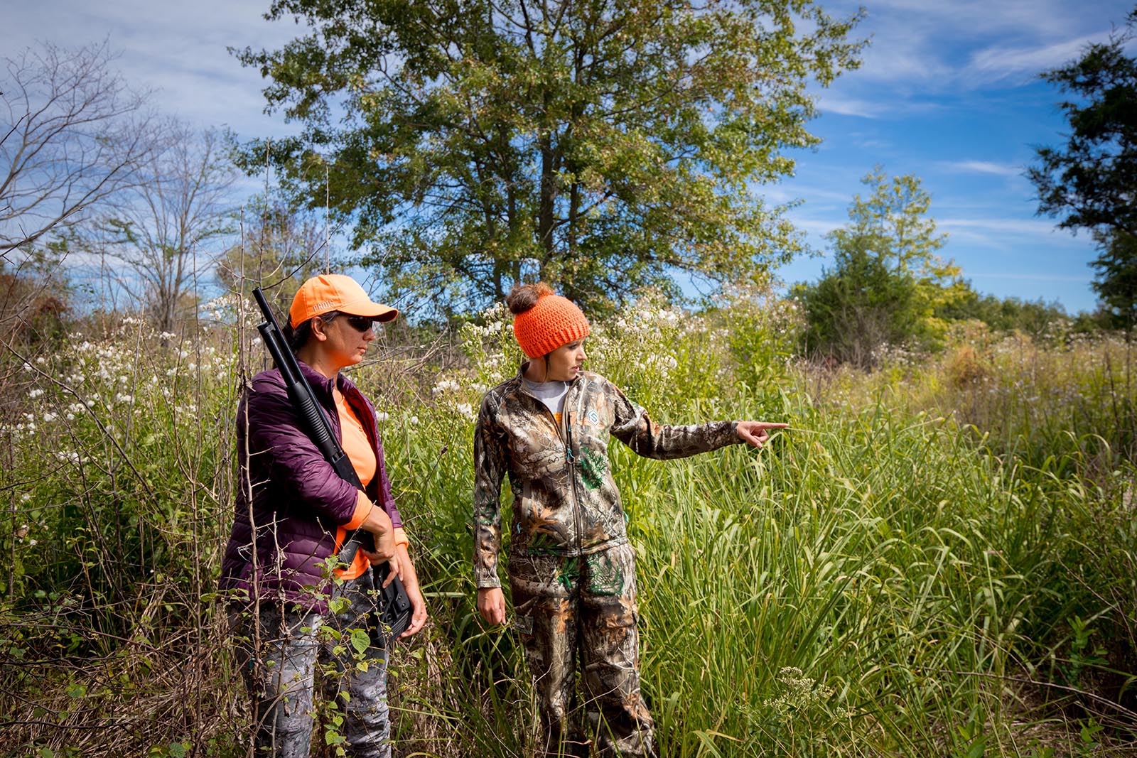 A photo of two women in camouflage and blaze orange standing in a field, with one woman directing the woman holding a gun.