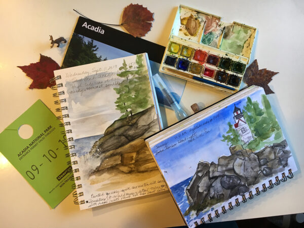 An image of a variety of journaling essentials such as a park pass and map as well as watercolors and a brush (in this case one that holds water for the painting) and a few notebooks