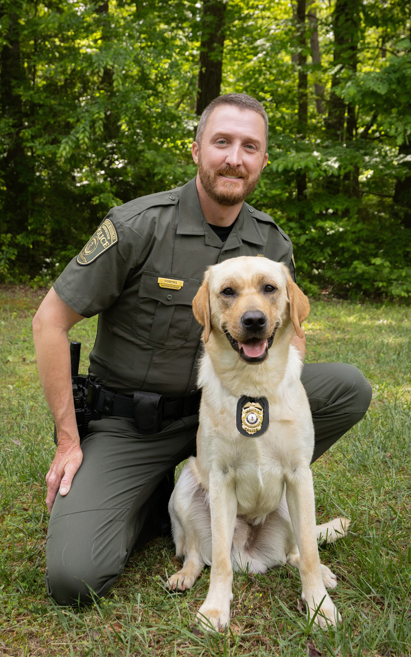 A photo of a uniformed law enforcement officer kneeling next to a dog with a badge on its collar.