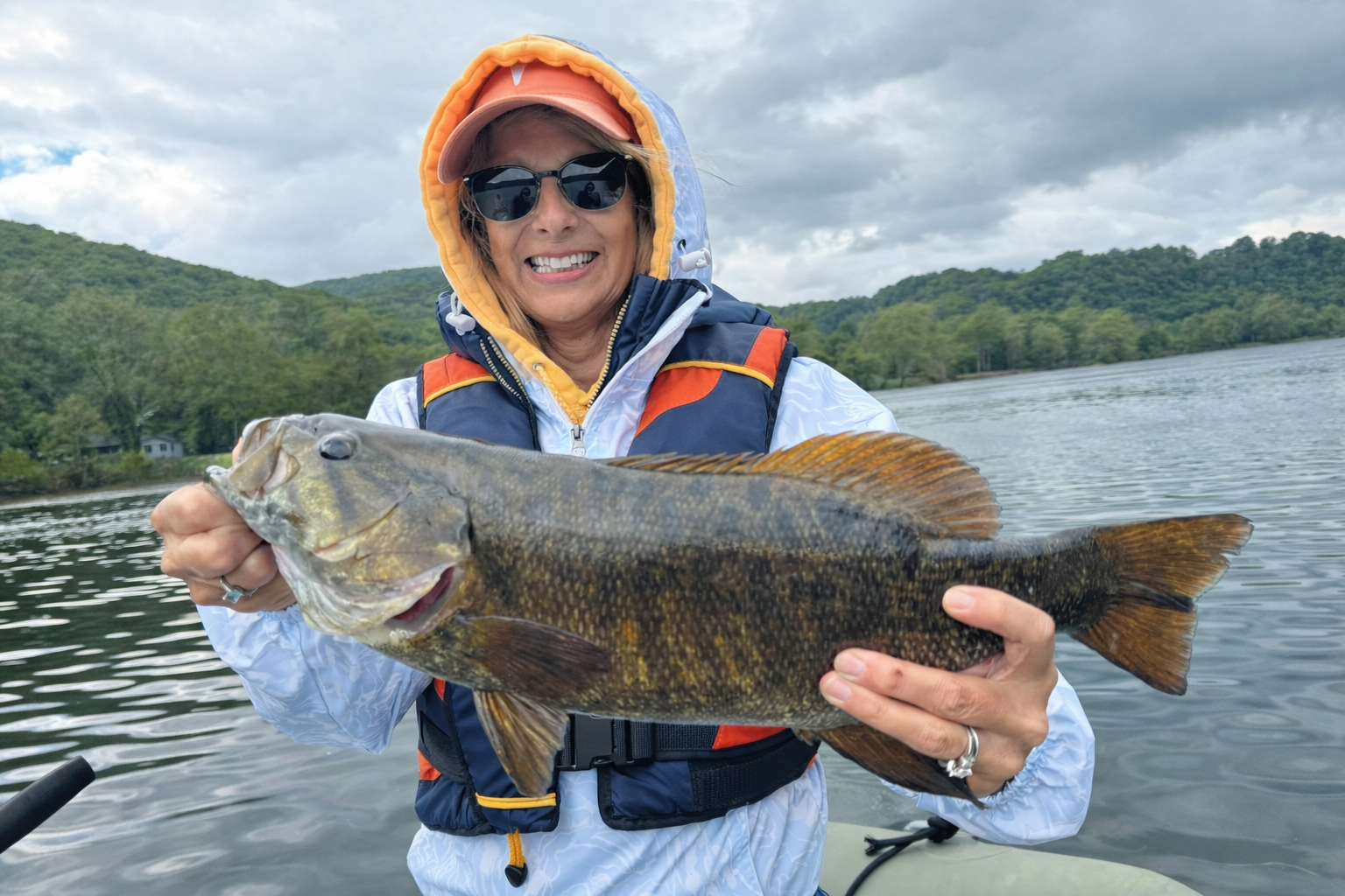 A photo of a woman smiling and holding a large smallmouth bass with a river in the background.