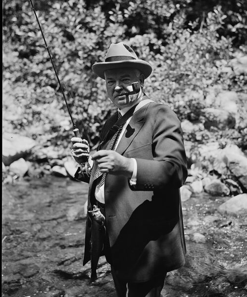 A black-and-white photo of President Herbert Hoover, dressed in a suit and with a pipe in his mouth, standing in a river holding a fly rod and a trout. 