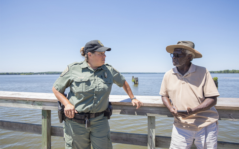 A photo of a female Conservation Police Officer in uniform speaking with a man standing on a pier.