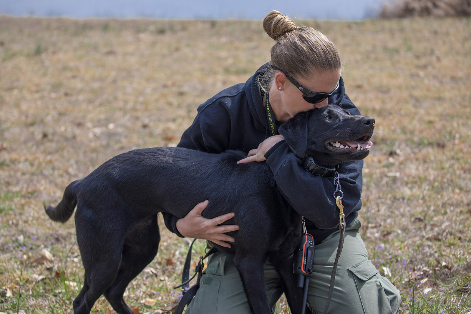 A photo of a female Conservation Police Officer kneeling and kissing a black lab dog on the head.