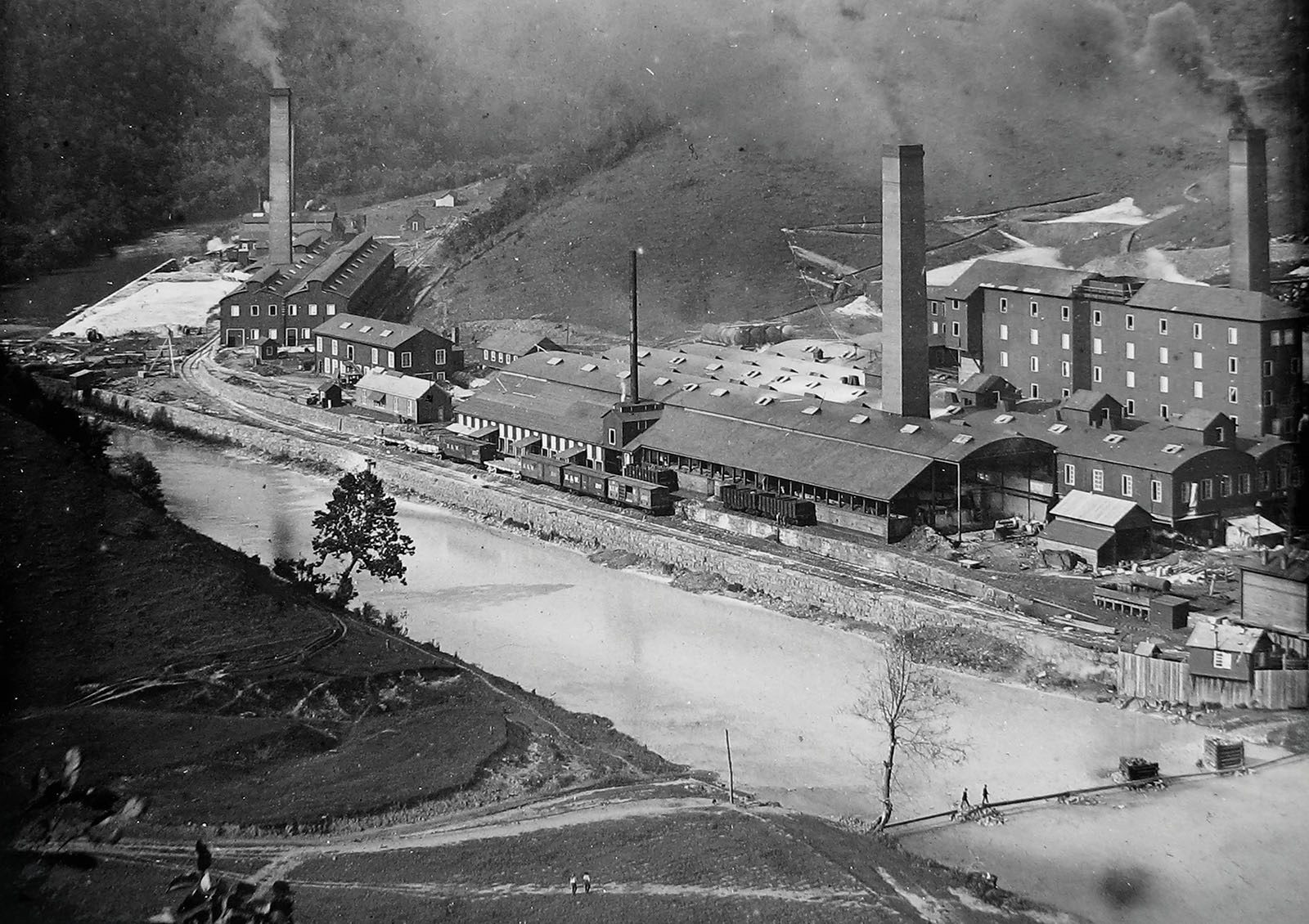 An old, black-and-white photo of a large chemical plant on the banks of a river, with smokestacks spewing dark smoke into the air.