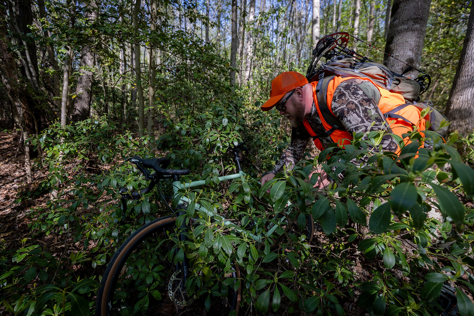 A photo of a man in camouflage and blaze orange covering a bike with bushes in the woods. 