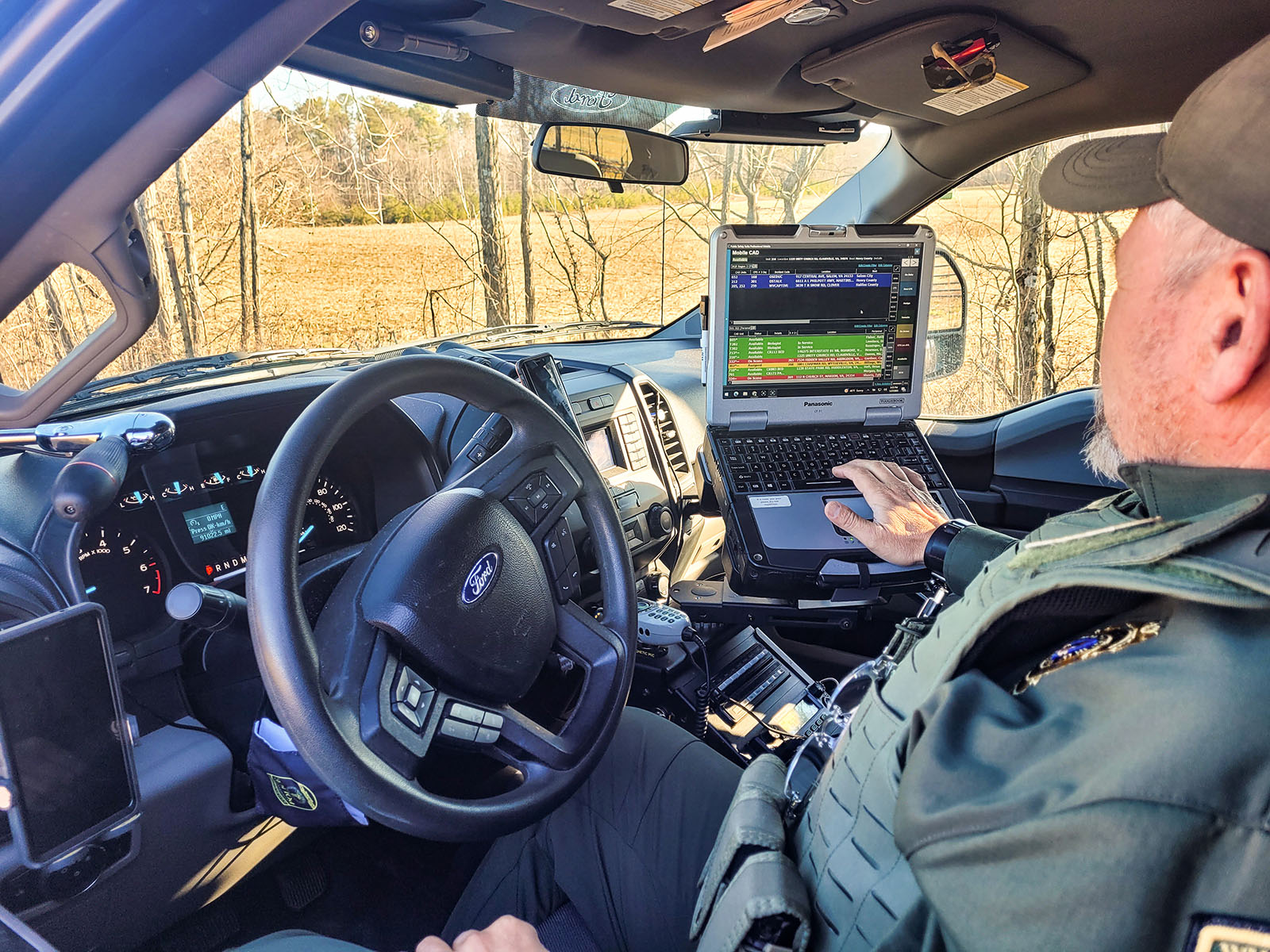 A photo of a Conservation Police Officer sitting in the driver's seat of his truck while looking at a laptop computer screen mounted to the center console.