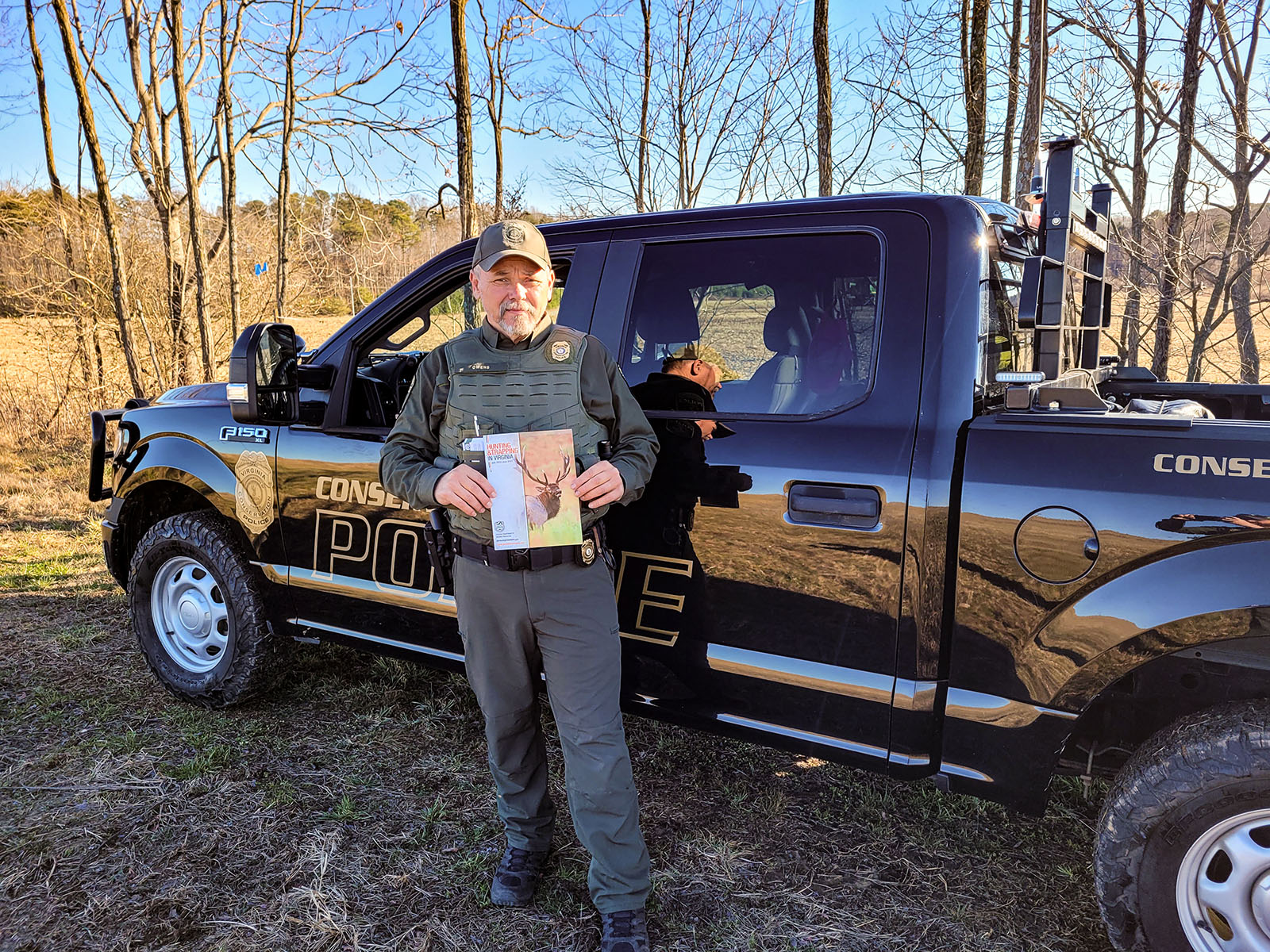 A photo of a Conservation Police Officer in uniform standing in front of his truck holding a printed copy of the 2023-24 Virginia Hunting and Trapping Regulations book.