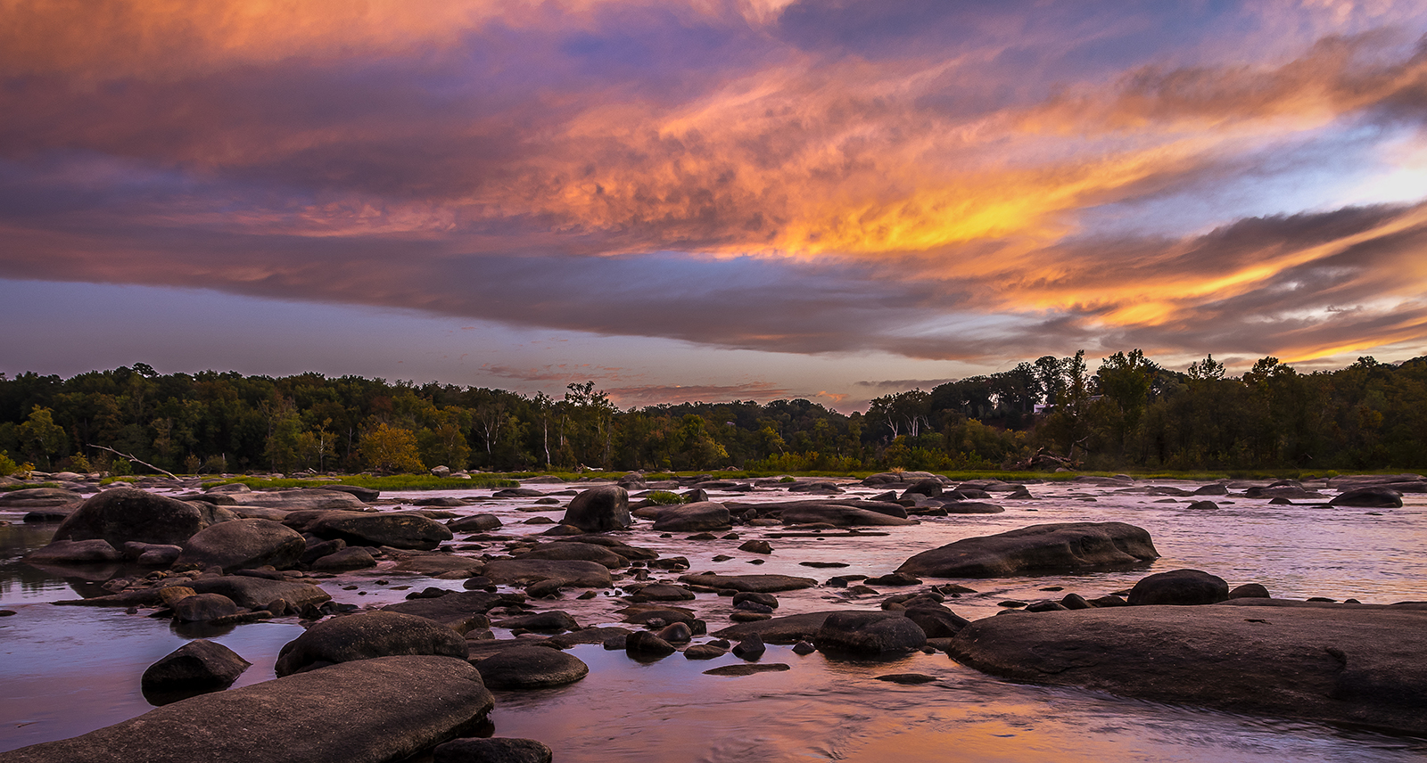 A photo of a gorgeous orange, blue, and pink sunrise sky above the water and rocks of Pony Pasture on the James River in Richmond.