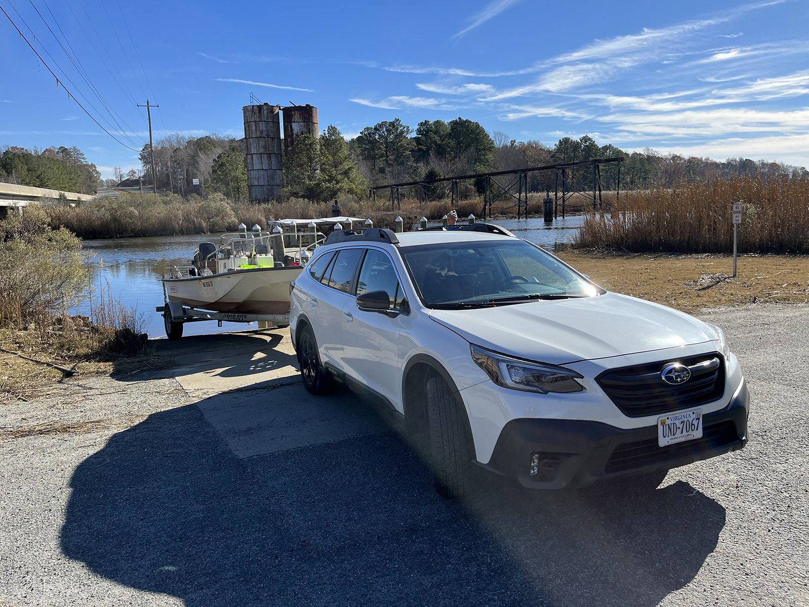 A white car pulling a small boat, backing down a boat ramp into a river.