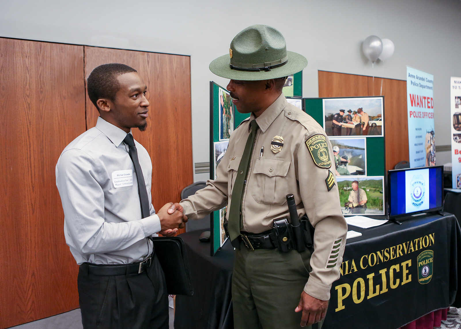 A photo of a man in uniform shaking the hand of a younger man in front of a table full of recruiting material.