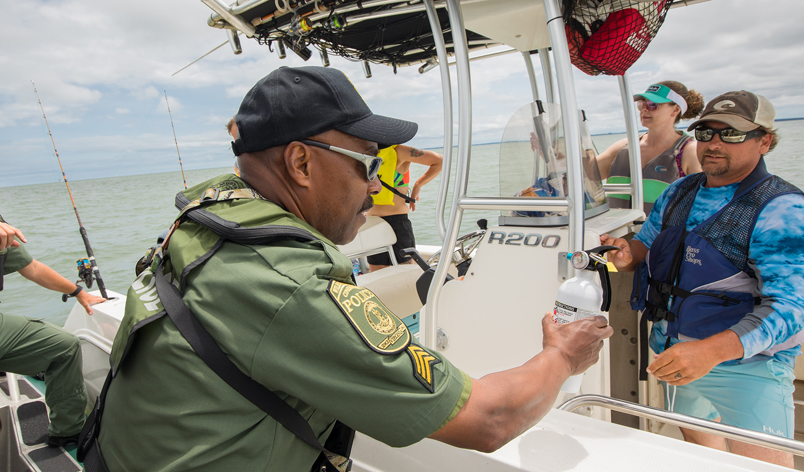 A photo of a man in uniform and on a boat holding a fire extinguisher as he speaks to two people on a boat.