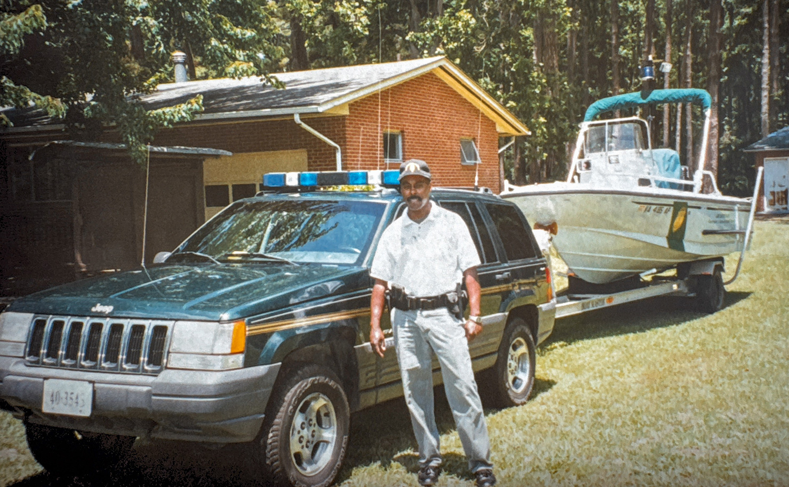 A photo of a man in uniform with a gun and badge standing in front of a Jeep hooked to a boat trailer.