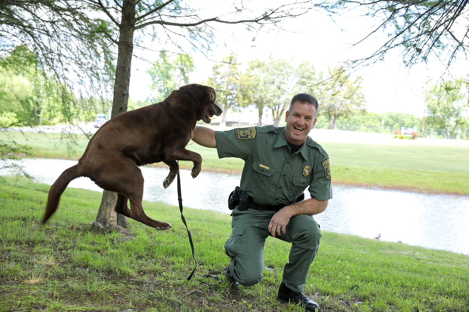 A photo of a brown Labrador Retriever jumping excitedly next to a kneeling Conservation Police Officer. 