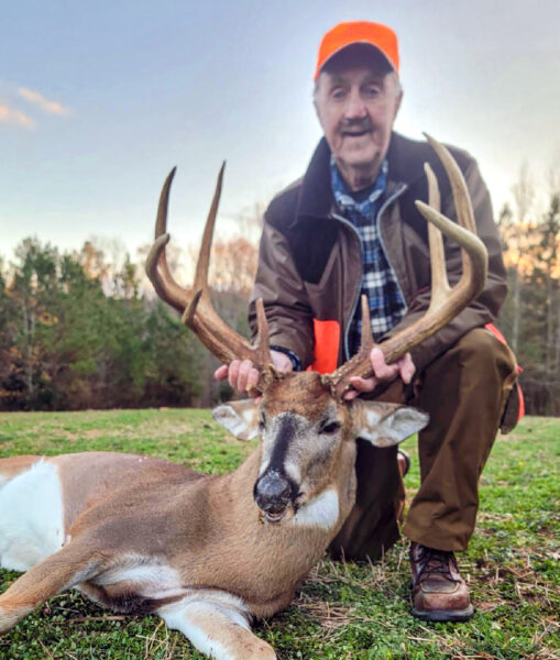A photo of an older man kneeling behind a large, antlered deer lying on the ground.