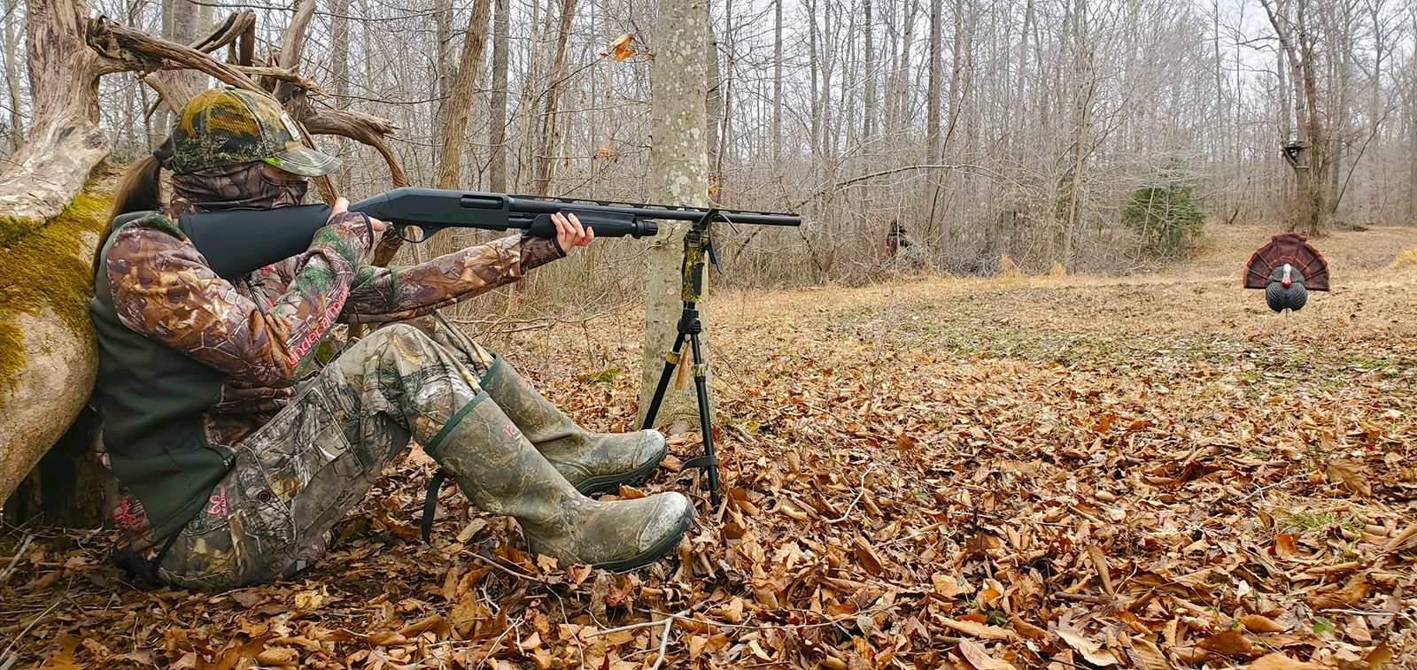 A photo of a young, female hunter in camouflage sitting on the ground against a tree, aiming a shotgun at a plastic turkey decoy.