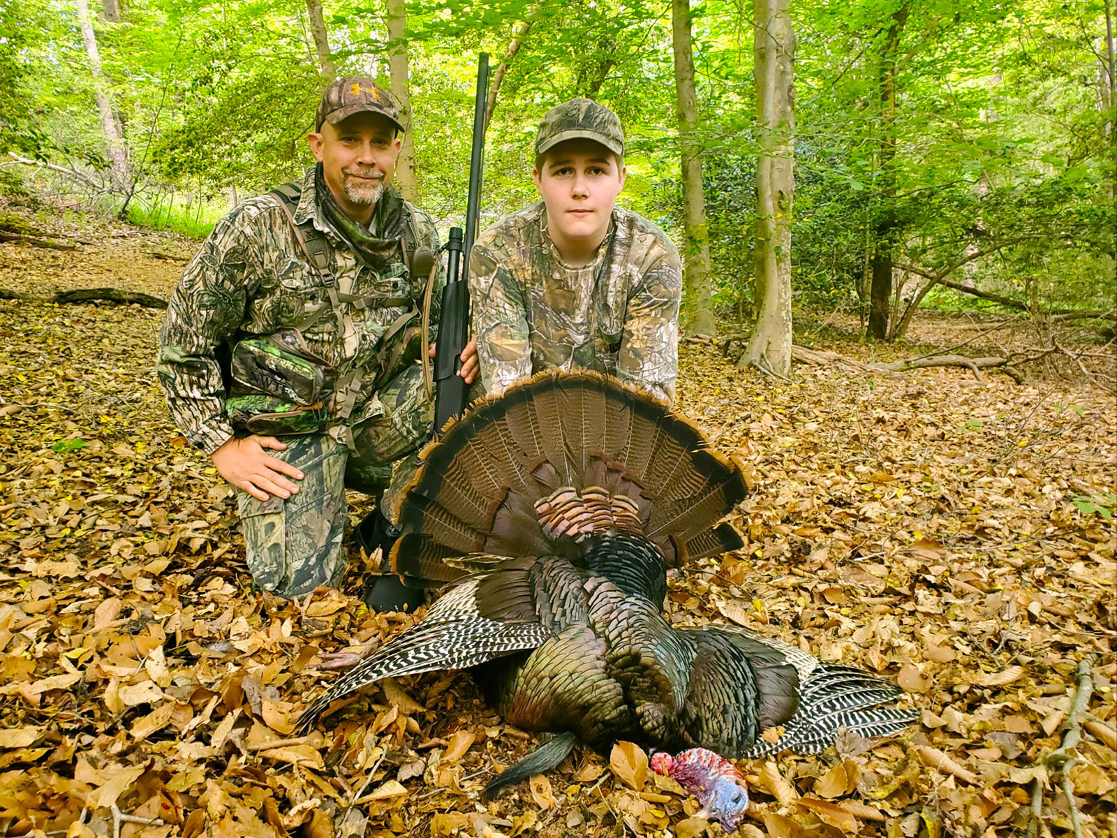 A man and a teenage kneeling in the woods, behind a harvested wild turkey. 
