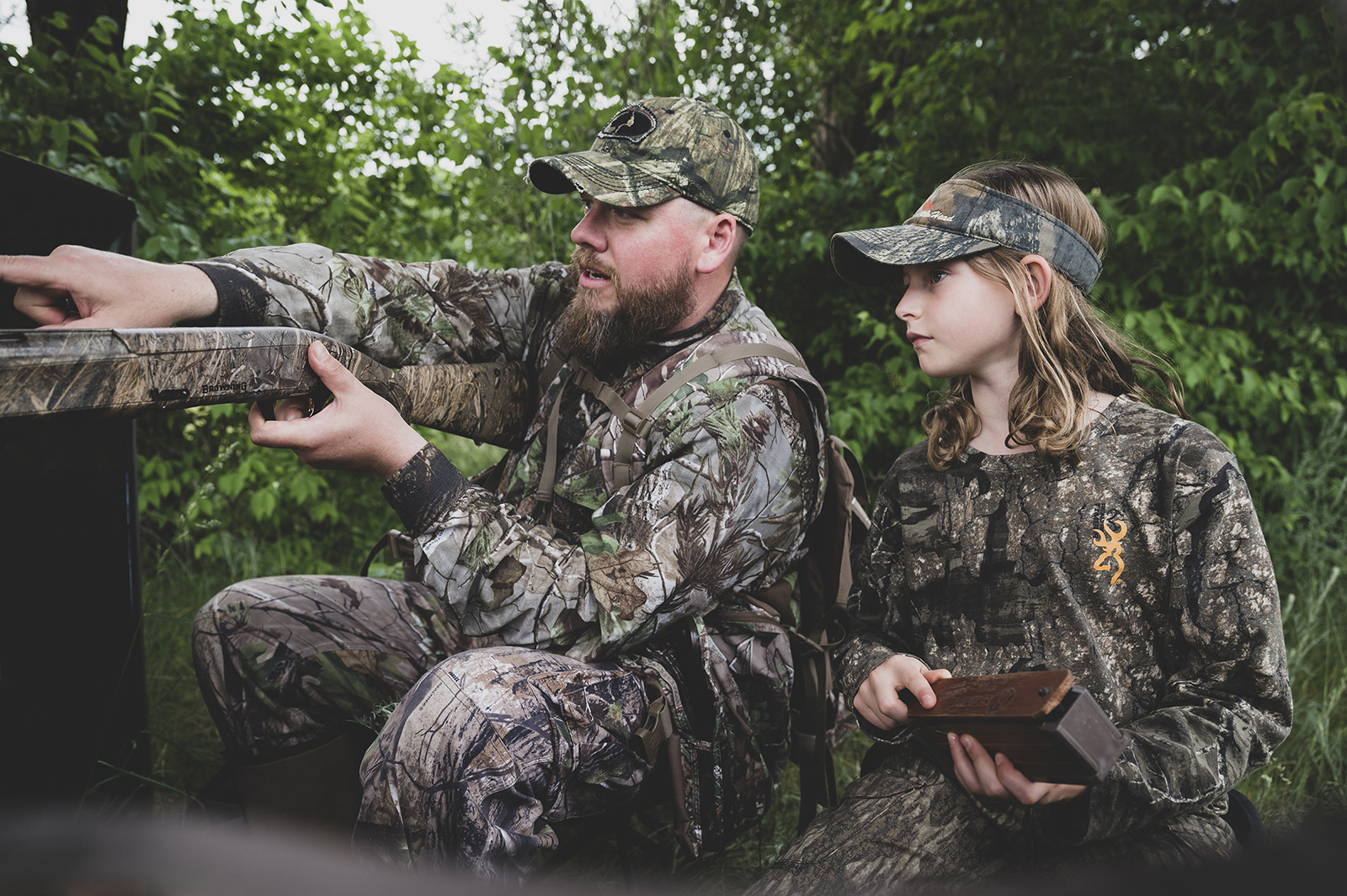 A photo of a male, adult hunter holding gun and pointing as a young, female hunter looks on and holds a turkey call. 