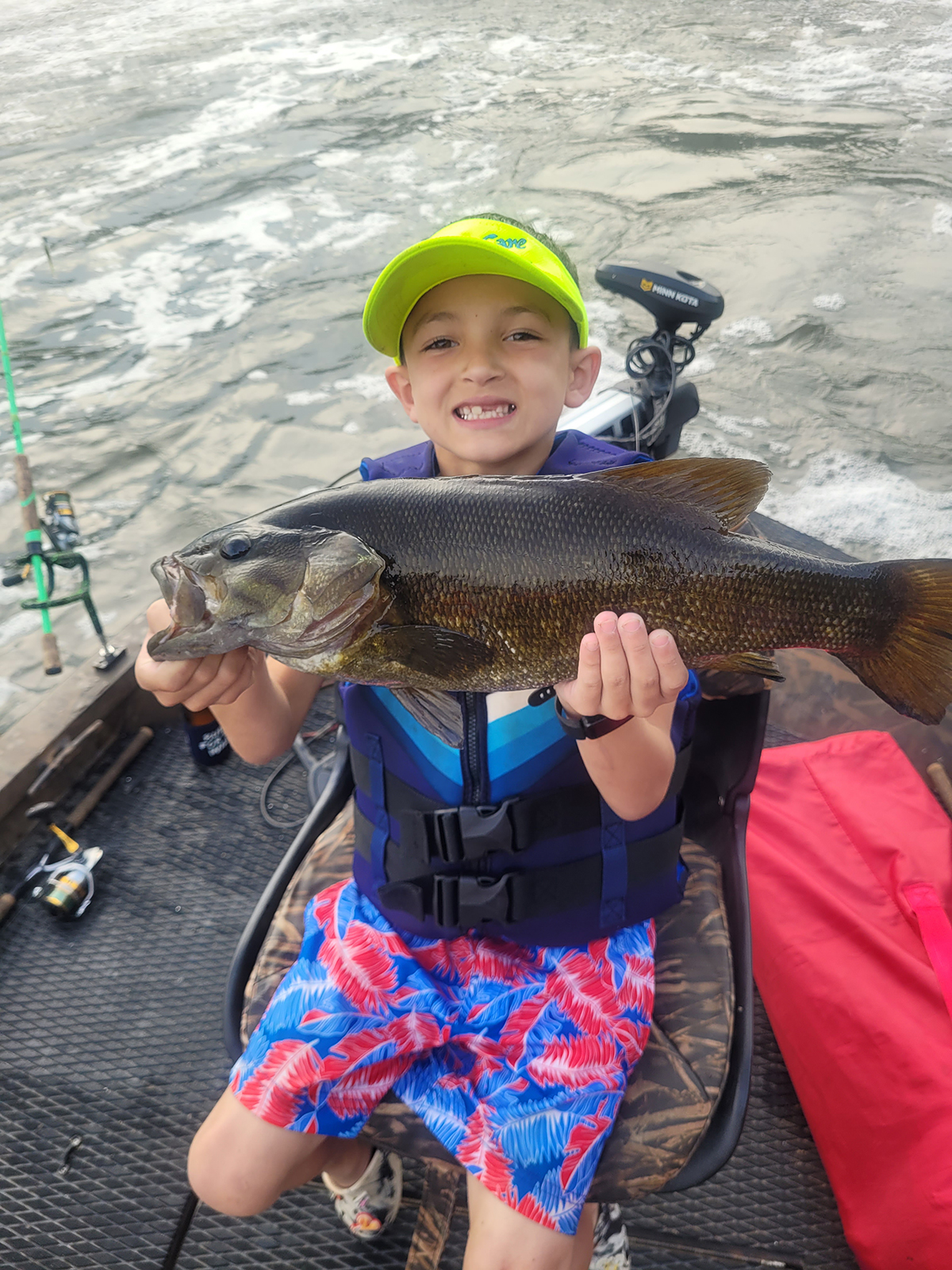 A photo of a young boy on a boat, wearing a life jacket, holding up a large smallmouth bass.