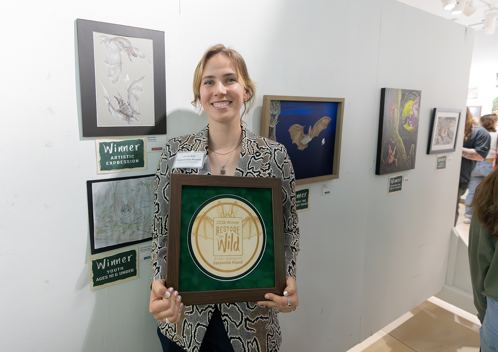 A photo of a woman holding a large winner plaque standing by a wall with art hanging on it. 