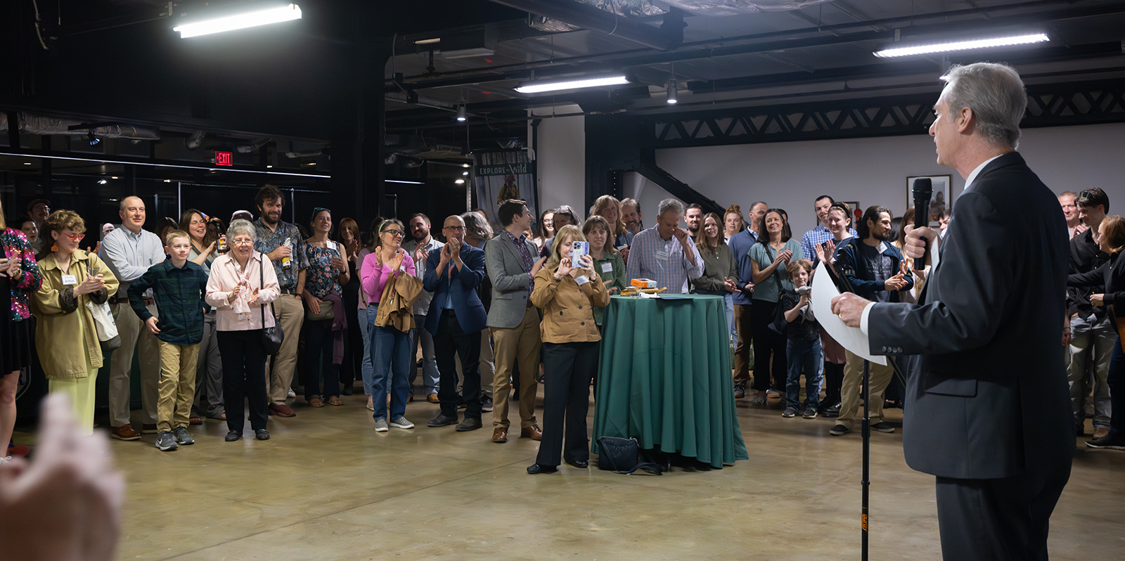 A photo of a man in a suit with a microphone addressing a crowded room.