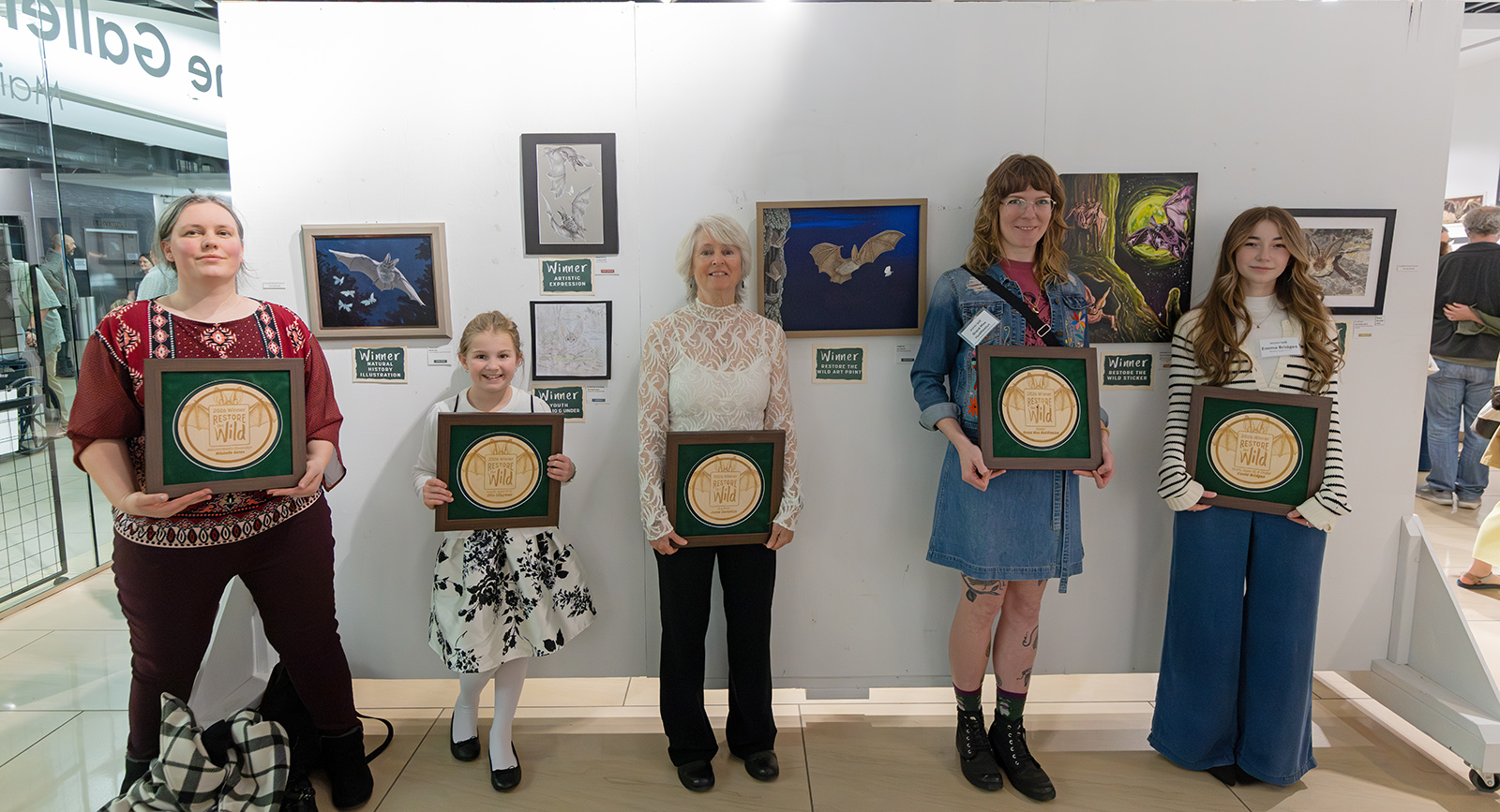 A photo of five people standing by a wall of art holding large winner plaques. 