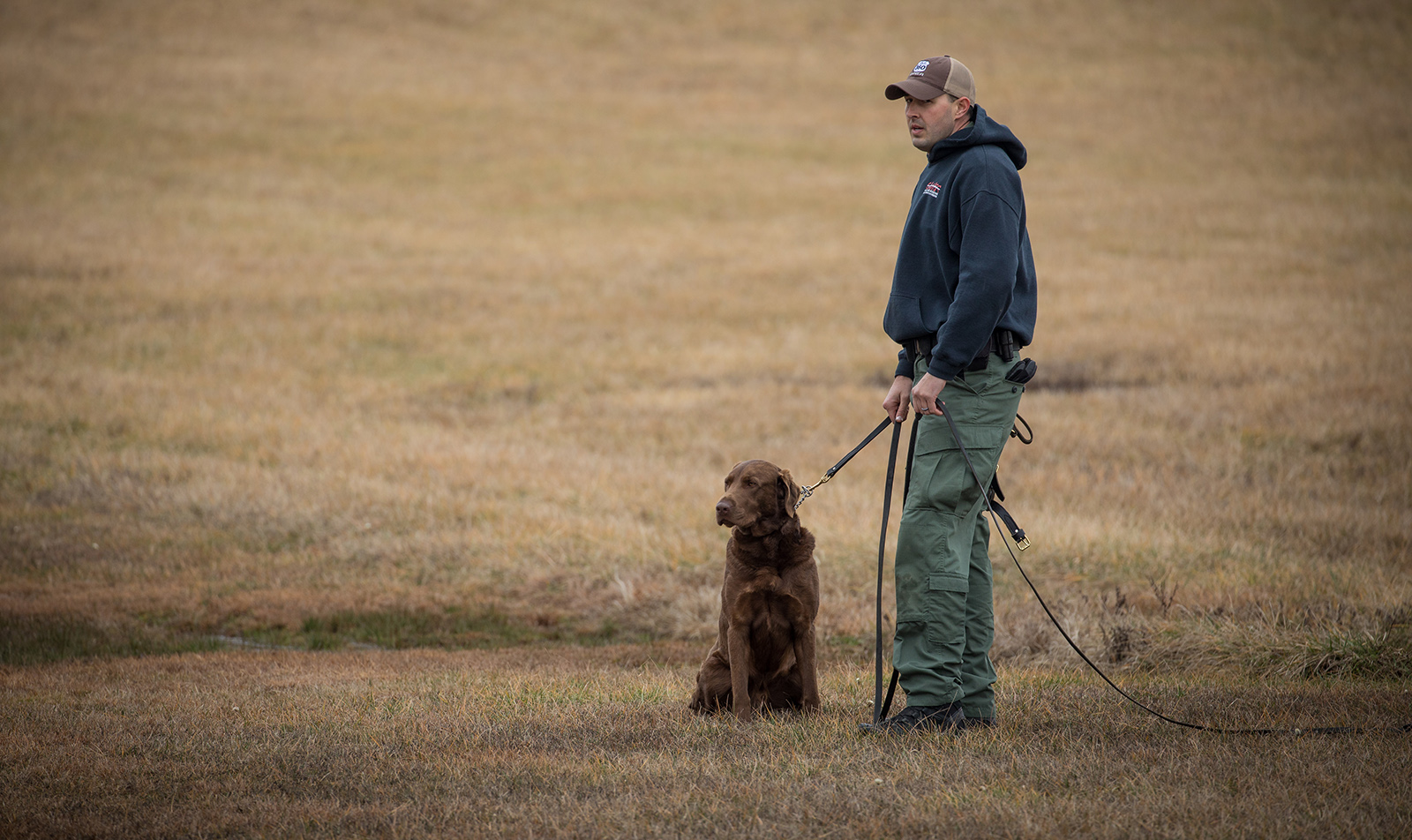 A photo of a man standing, holding the leash of a large brown dog that's sitting quietly.