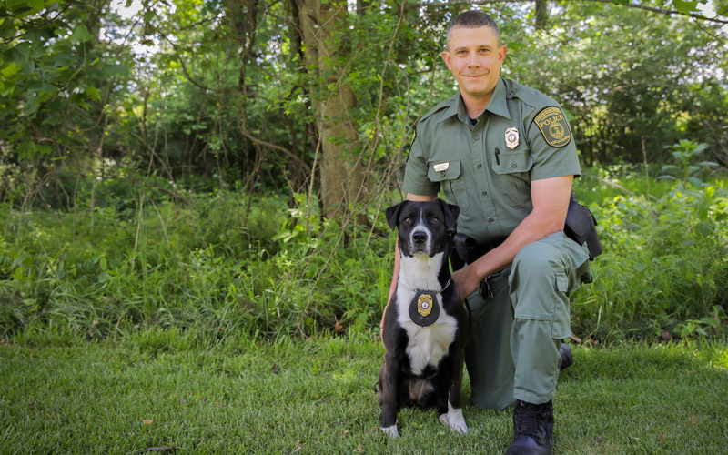 A photo of a man in law enforcement uniform kneeling next to a black and white dog with a badge around its neck.