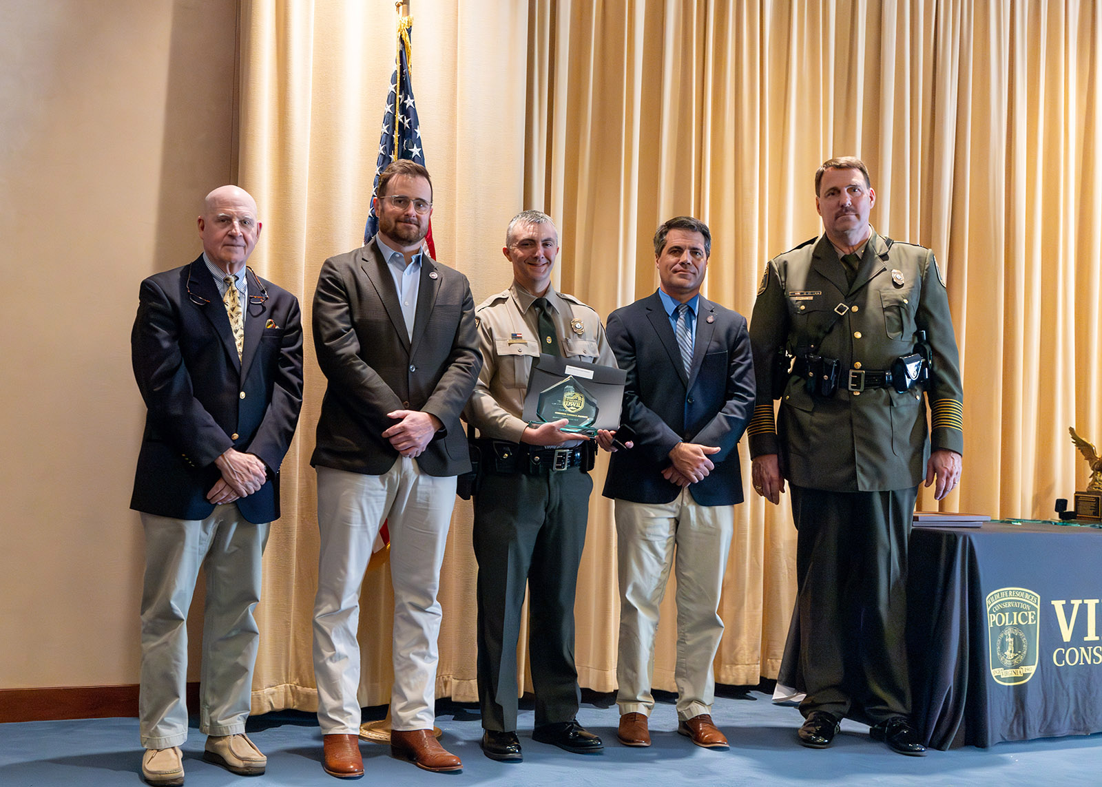 A photo of five men standing on a stage, with the man in the center in Conservation Police uniform and holding a plaque. 
